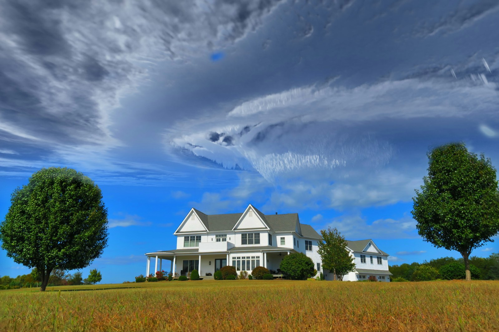white and gray house between two green tall trees under gray clouds forming swirl during daytime home